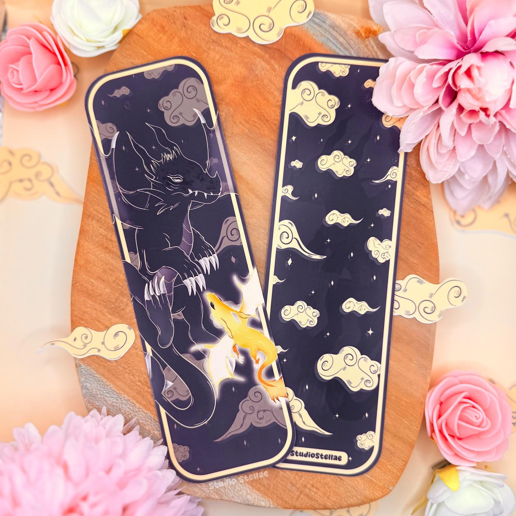 Dark fantasy dragon bookmark set displayed on a warm wooden board surrounded by soft pink flowers and decorative cloud accents. Two uniquely shaped laminated bookmarks are shown side by side, featuring a midnight sky background with swirling clouds, a powerful shadow dragon silhouette, and glowing golden dragon fire. The dramatic black and gold color contrast highlights the romantasy aesthetic inspired by Fourth Wing, perfect for dragon lovers and fantasy readers.