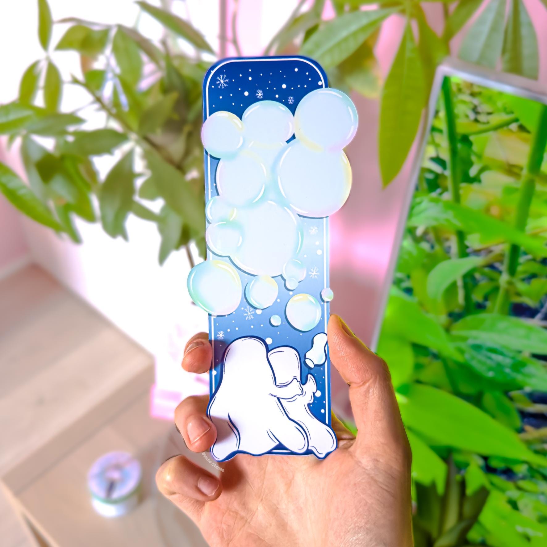 Close-up of the Ghost Couple Blowing Bubbles Bookmark held upright in front of leafy green plants in natural light, showing the back side of the design. The reverse features a dreamy starry night sky pattern filled with soft translucent blue bubbles and small sparkles, matching the pastel spooky winter aesthetic. The slim rectangular shape, rounded corners, and smooth matte laminated finish are clearly visible, showing the double-sided waterproof page marker quality.