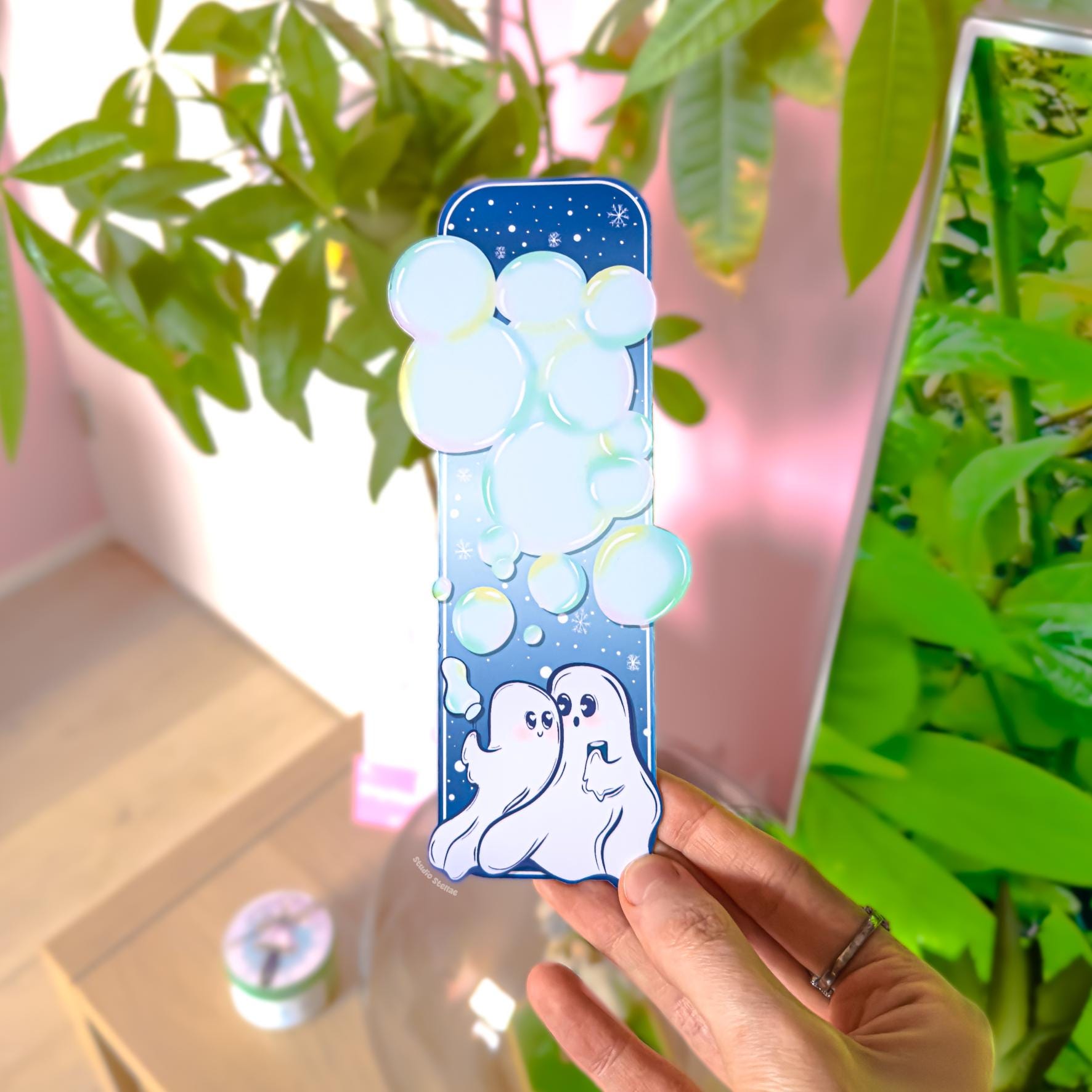 Close-up of the Ghost Couple Blowing Bubbles Bookmark held in front of leafy green plants in natural light. The pastel blue night sky, soft glowing bubbles, and two white ghosts at the bottom are clearly visible. The uniquely shaped bottom edge and matte laminated finish can be seen in detail. This lifestyle image demonstrates how the waterproof Halloween and Valentine bookmark looks during everyday reading.