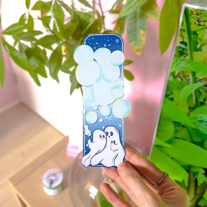 Close-up of the Ghost Couple Blowing Bubbles Bookmark held in front of leafy green plants in natural light. The pastel blue night sky, soft glowing bubbles, and two white ghosts at the bottom are clearly visible. The uniquely shaped bottom edge and matte laminated finish can be seen in detail. This lifestyle image demonstrates how the waterproof Halloween and Valentine bookmark looks during everyday reading.