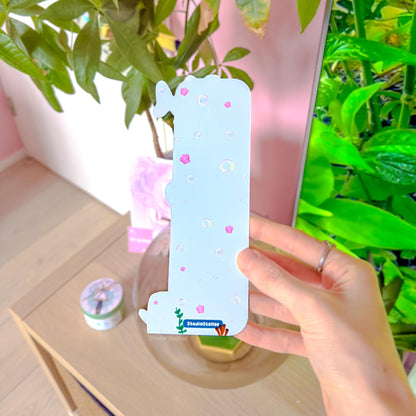 Close-up of the ocean animal bookmark held in front of leafy green plants in natural light. The front design features chibi sharks, orange octopus, and squid swimming in pastel blue water with tiny bubbles and sea details. The uniquely shaped bottom edge and smooth matte laminated surface are visible, showing durability and vibrant color.