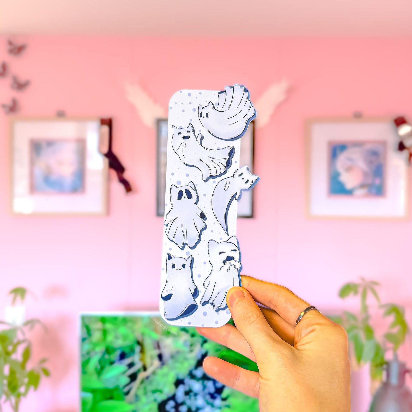 Close-up of a hand holding the Witchy Cat Ghost Bookmark in a pink room setting with framed wall art in the background. The front shows layered ghost cat shapes peeking past the edges, floating on a soft pastel purple base with small dot details. Matte laminated finish and die-cut silhouette are visible, making this a cute spooky bookmark and Halloween kitty page marker for readers.