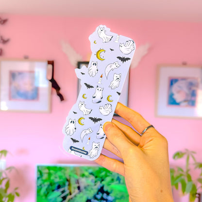 Close-up of a hand holding the Witchy Cat Ghost Bookmark in the same pink room setting, now showing the back design. The reverse features a repeating pastel purple pattern with tiny ghost cats, spooky bats, crescent moons, and candle details like a Halloween night scene. The smooth matte lamination, rounded corners, and sturdy thickness are visible, highlighting this double-sided spooky reading accessory as a bookish reader gift.