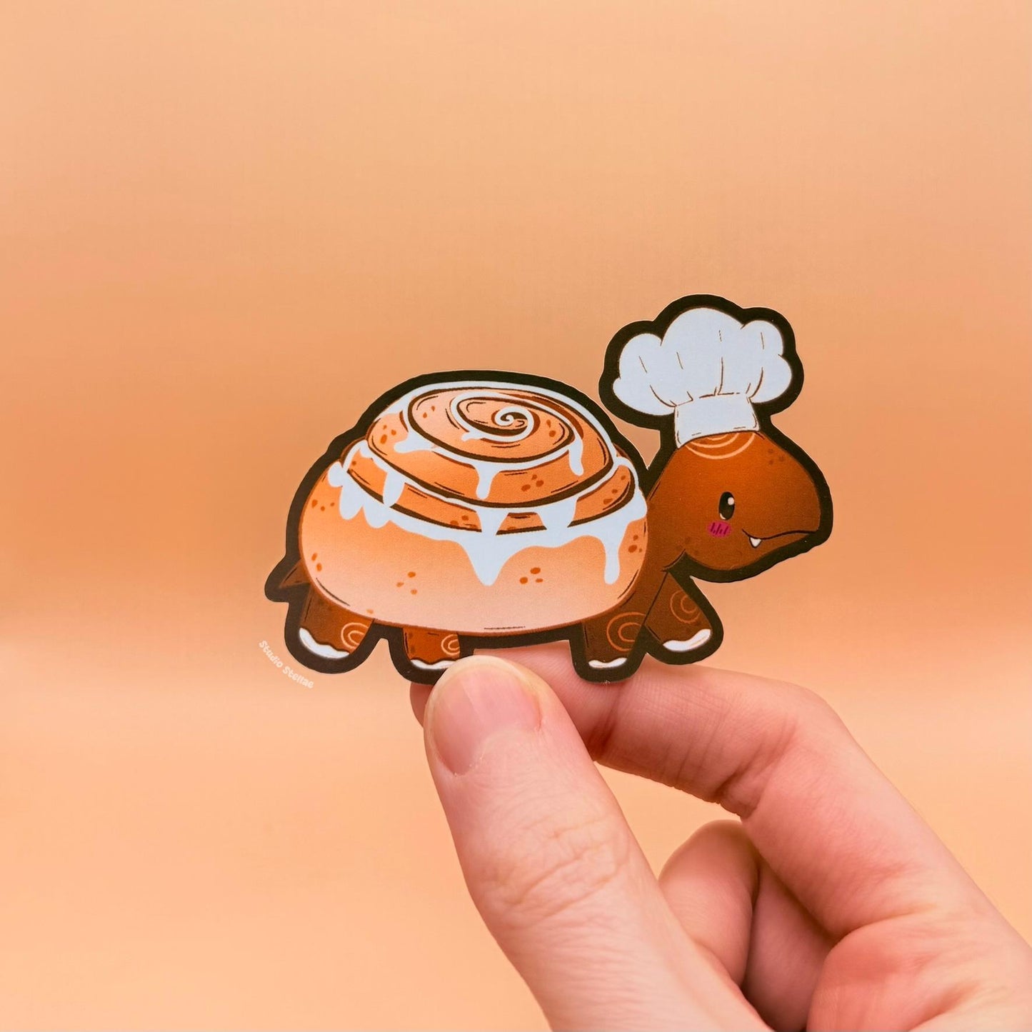 Close-up of a hand holding and peeling the cinnamon roll turtle sticker to show size, flexibility and detail. The matte laminated surface and clean die cut edges highlight the durable finish and soft texture. This cute pastry turtle decal applies easily to journals, planners, laptops and water bottles, making it a fun and cozy comfort food decoration for stationery lovers and bakery fans.