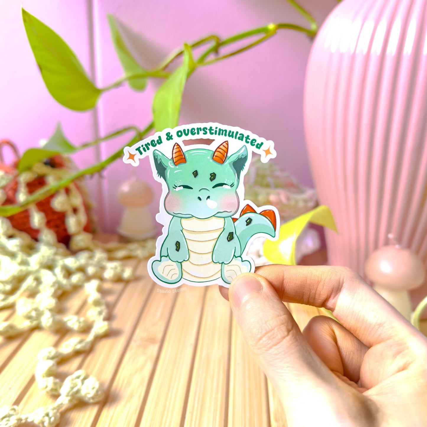 Close-up of a hand holding the tired baby dinosaur sticker near green plants in soft natural light to show true size and color. The sleepy kawaii dino expression highlights relatable overstimulation and burnout humor. This matte laminated vinyl decal works perfectly on laptops, planners, journals and water bottles as a gentle mental health reminder.