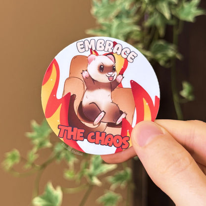 Close-up lifestyle photo of a hand holding the Embrace the Chaos ferret sticker near green plant leaves, showing the sticker’s true size and color in natural light. The cute kawaii ferret sits on a chair surrounded by flames with the phrase “Embrace the Chaos,” mixing adorable animal art with funny chaotic humor. Handmade matte laminated decal for laptops, planners, journals and water bottles, perfect for ferret lovers and chaos enjoyers.
