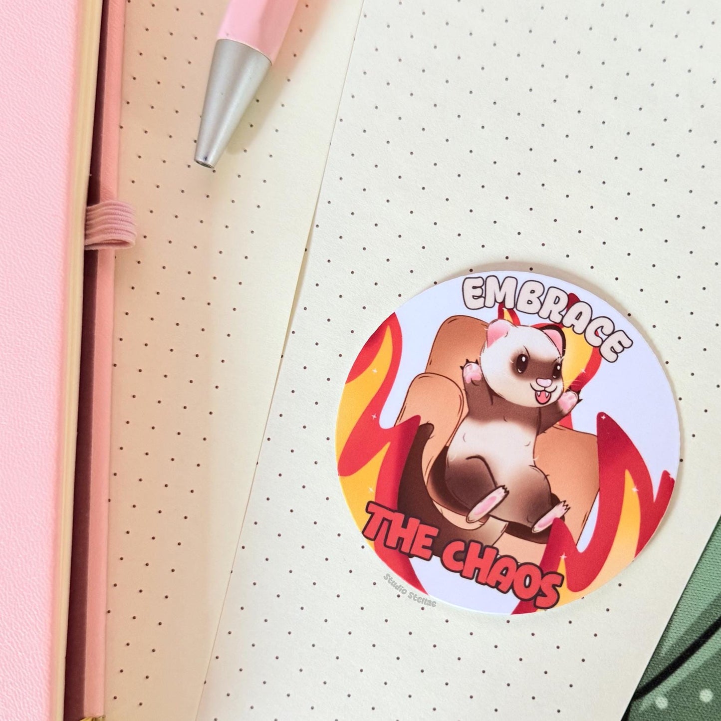 The Embrace the Chaos ferret sticker displayed on a notebook or planner page in a cozy workspace, showing how the design looks in everyday stationery setups. The flames and playful ferret add humorous chaotic energy to journals and study notes. This funny animal decal works perfectly for planners, laptops and water bottles.