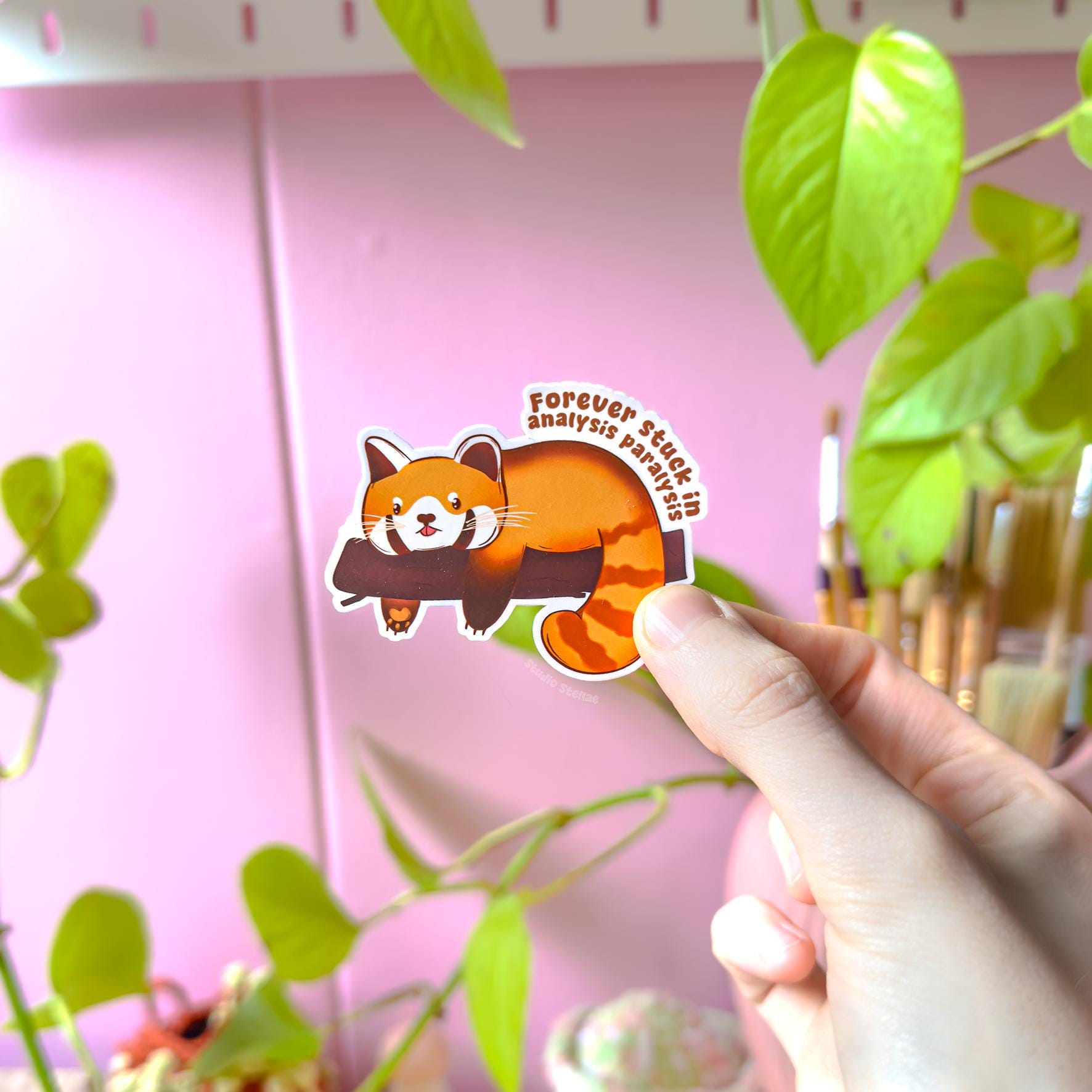 Close-up of a hand peeling and placing the red panda sticker onto a smooth surface near a small plant to show scale and flexibility. The matte laminated finish and clean die cut edges highlight the durable, water resistant quality. This cute animal Analysis Paralysis sticker is easy to apply to journals, planners, water bottles or laptops, making it a fun and relatable overthinking humor decoration.