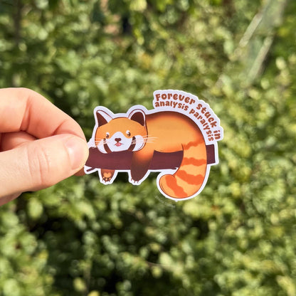 Lifestyle photo of the Analysis Paralysis red panda sticker held outdoors against soft green foliage, showing size and detail in natural light. The lounging kawaii animal design feels cozy and relatable, representing burnout, anxious thoughts and overthinking humor. This handmade matte vinyl decal works beautifully as a journal sticker, planner accessory or laptop decoration for ADHD and neurodivergent stationery fans.