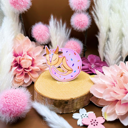 Close-up lifestyle photo of the frosted sugar cookie dragon sticker styled among pink flowers and soft decor to highlight its size and pastel colors. The tiny curled dragon pastry looks sweet and whimsical, blending cute food art with fantasy creature charm. This matte laminated decal works beautifully on journals, planners, laptops and water bottles as a cozy decorative sticker.