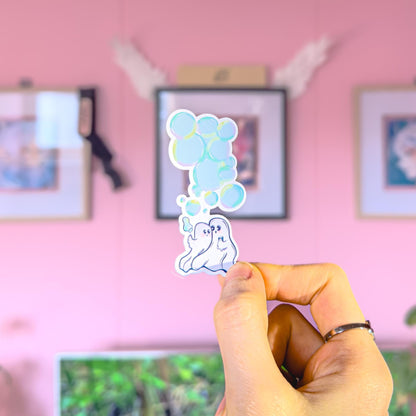 Close-up of a hand holding and applying the ghost couple sticker in a cozy pastel room, showing the size and flexibility of the matte laminated finish. The kawaii ghosts and bubbles stand out clearly, making it easy to decorate planners, journals, laptops or water bottles. This romantic spooky cute decal works beautifully as a small everyday reminder of love and comfort.