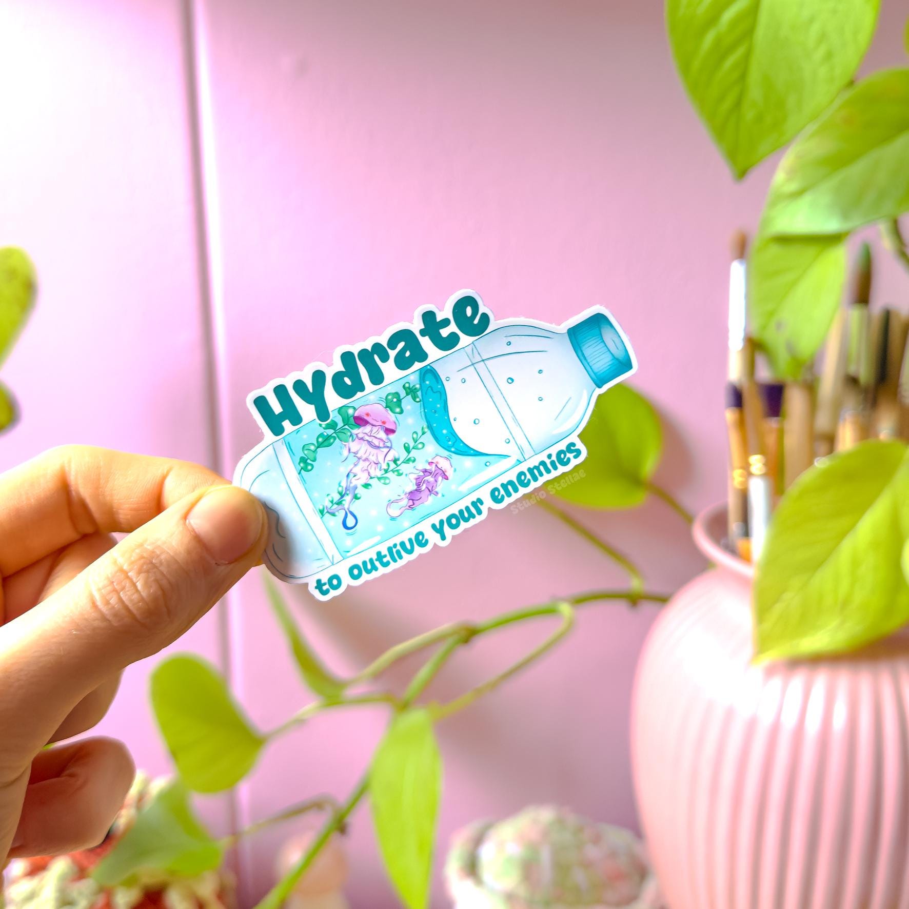 Close-up of a hand peeling and placing the jellyfish sticker onto a smooth surface near a plant to show size and flexibility. The matte laminated surface and clean die cut edges highlight the durable finish. This cute ocean animal decal applies easily to water bottles, planners, laptops and journals, making it a funny everyday hydration and mental health reminder.