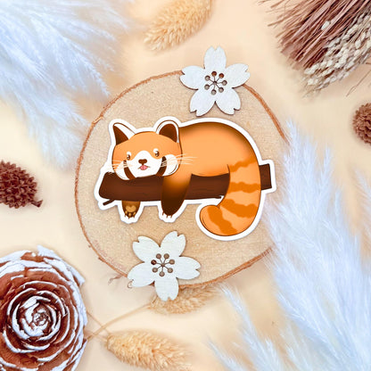 Sleepy red panda sticker arranged on a soft cream background with small florals and natural elements, creating a calm, cozy mood that matches the design. The relaxed pose and matte surface are clearly visible, emphasizing texture and print quality. This gentle flat lay demonstrates how the lazy panda fits beautifully into planners, journals or aesthetic desk setups.
