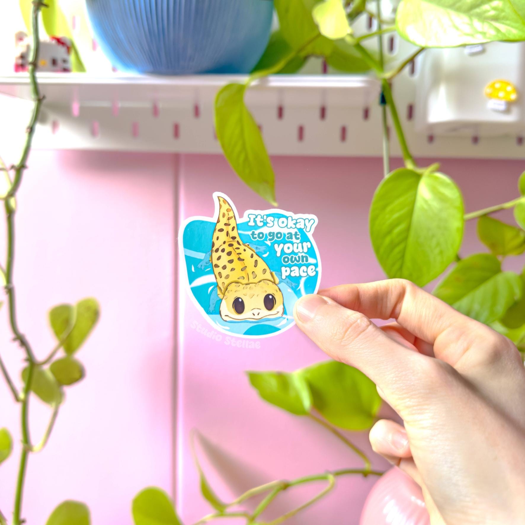 Close-up of a hand peeling and placing the gecko sticker onto a smooth surface near a plant to show size and flexibility. The matte laminated surface and clean die cut edges highlight the durable finish. This cute yellow lizard decal applies easily to journals, planners, laptops and water bottles, making it a calming everyday reminder to slow down and go at your own pace.