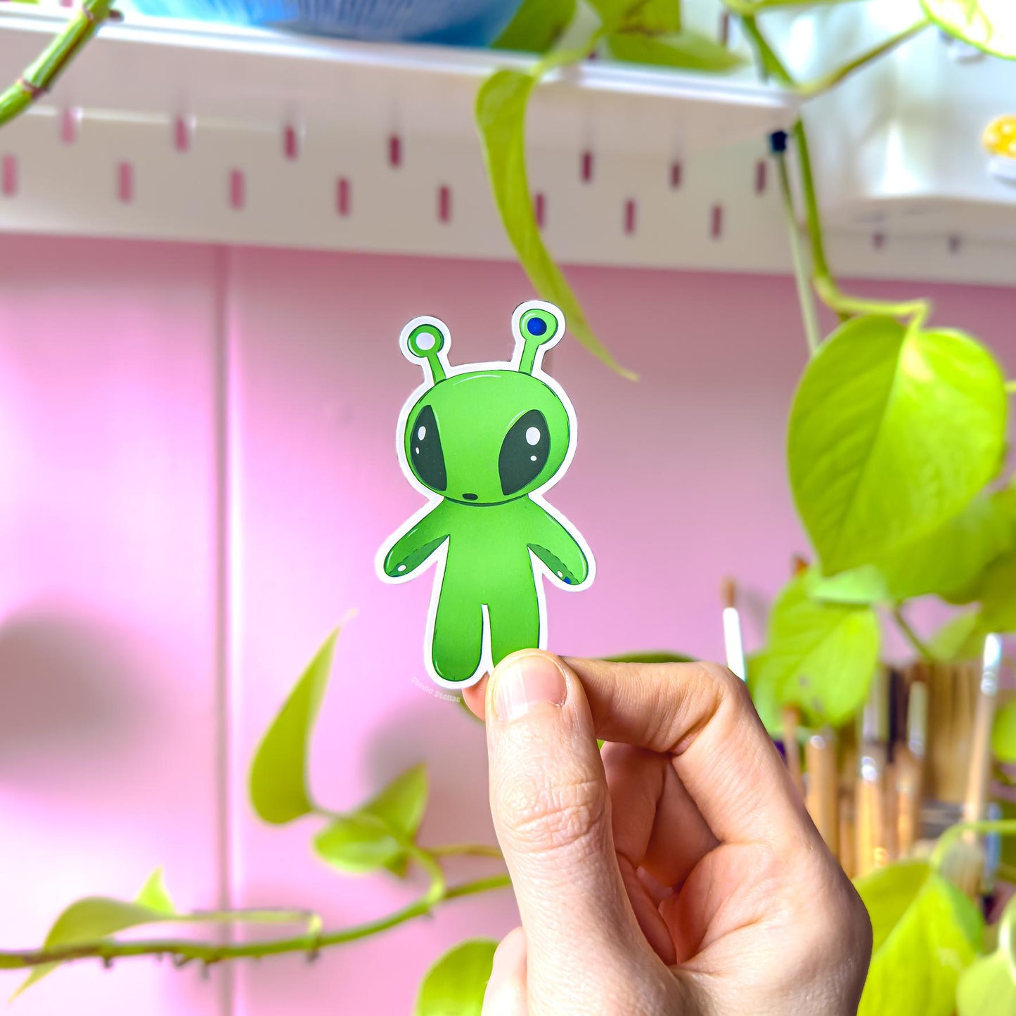 Close-up of a small rounded Aftonsparv alien sticker pinched between fingers to show scale and detail. The compact kawaii design features big eyes and soft pastel tones with a durable matte laminate surface. Perfect for smaller spaces like planners, journals, pencil cases or water bottles, and a sweet mini accent from the IKEA alien fan art sticker set.