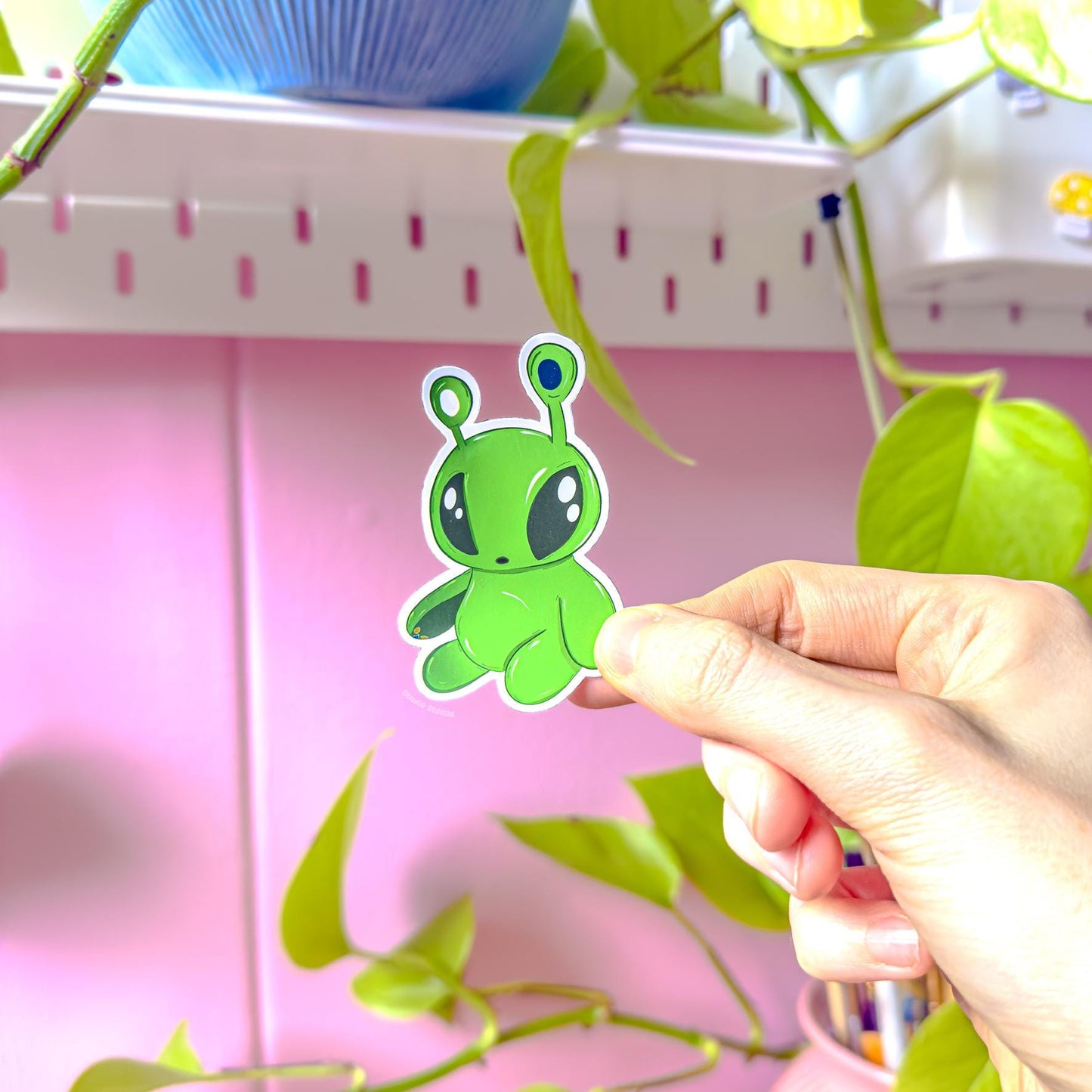 Hand holding the taller Aftonsparv alien character sticker in soft daylight, highlighting its elongated chibi shape, bold green color and clean edges. This plushie inspired kawaii alien decal shows the sticker’s true size and texture, making it easy to picture on laptops, planners, phone cases or water bottles. A playful addition to any cute stationery or tech setup.
