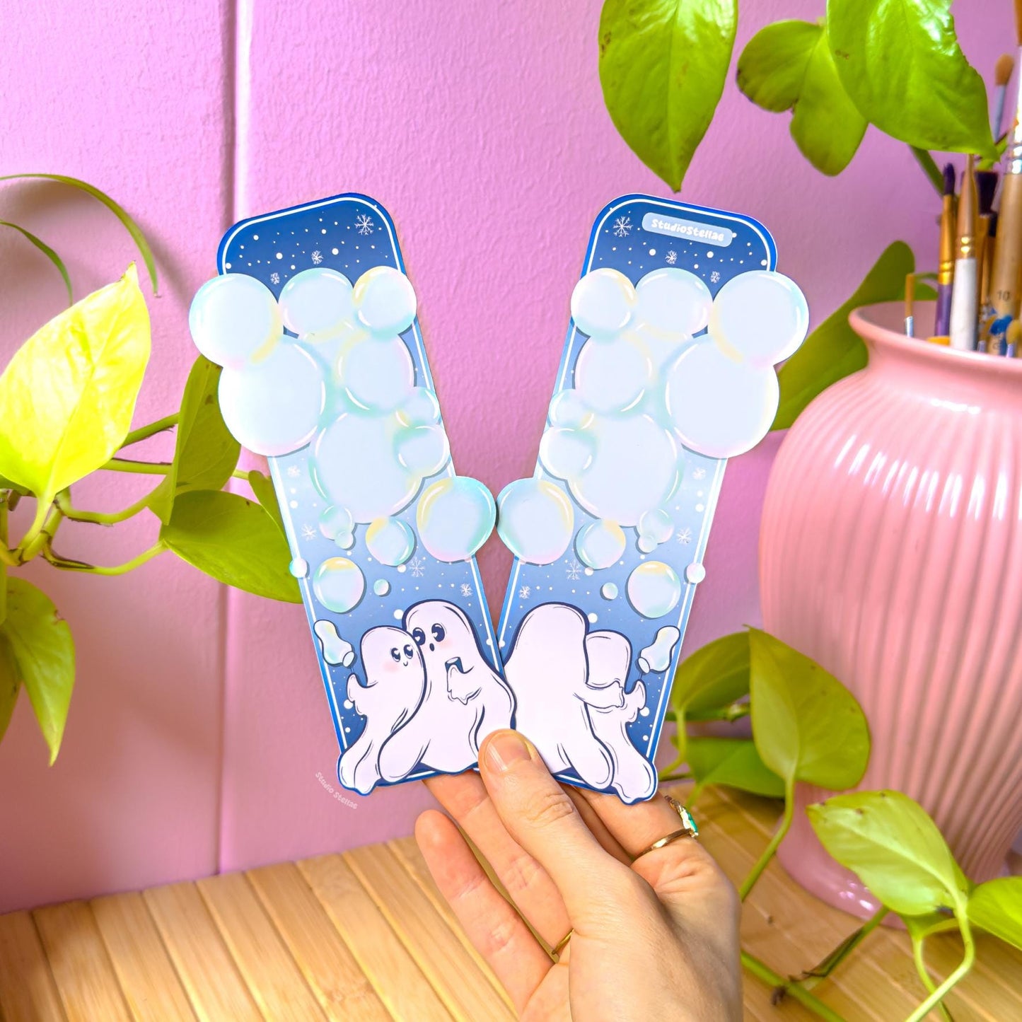Hand holding the pastel ghost couple bookmark in a cozy workspace with a pink wall, leafy green plant, and soft pastel decor. The waterproof laminated page marker shows two cute ghosts facing each other while blowing bubbles into a starry blue sky. The image demonstrates real-life scale and how the romantic spooky bookmark looks in hand, ideal for book lovers, readers, and anyone who enjoys pastel Halloween aesthetics.