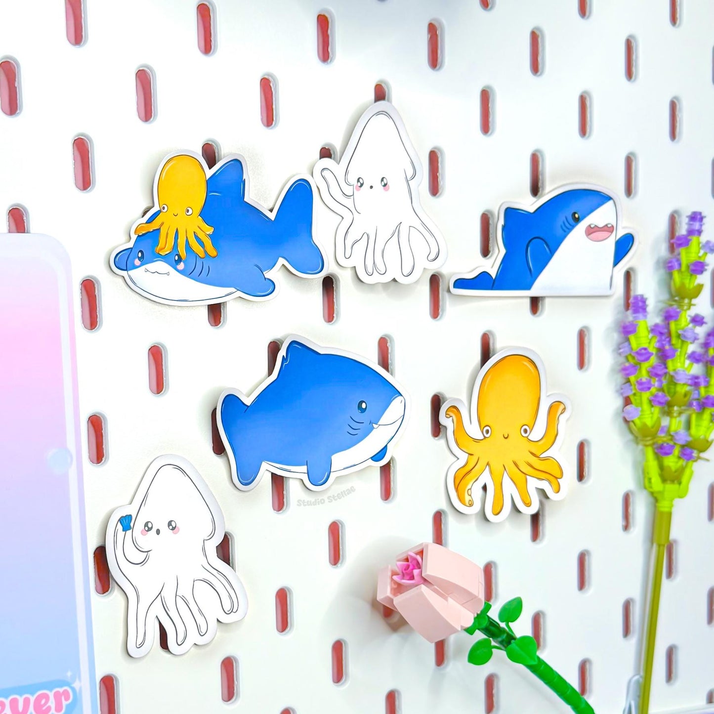 Ocean animal stickers arranged neatly on a pegboard style wall background, clearly showing each individual shark, squid and octopus design side by side. This layout highlights the variety and character expressions included in the kawaii sticker pack along with crisp die cut edges and bold colors. A waterproof sea life decal set ideal for planners, laptops, journals and fun stationery decorating.