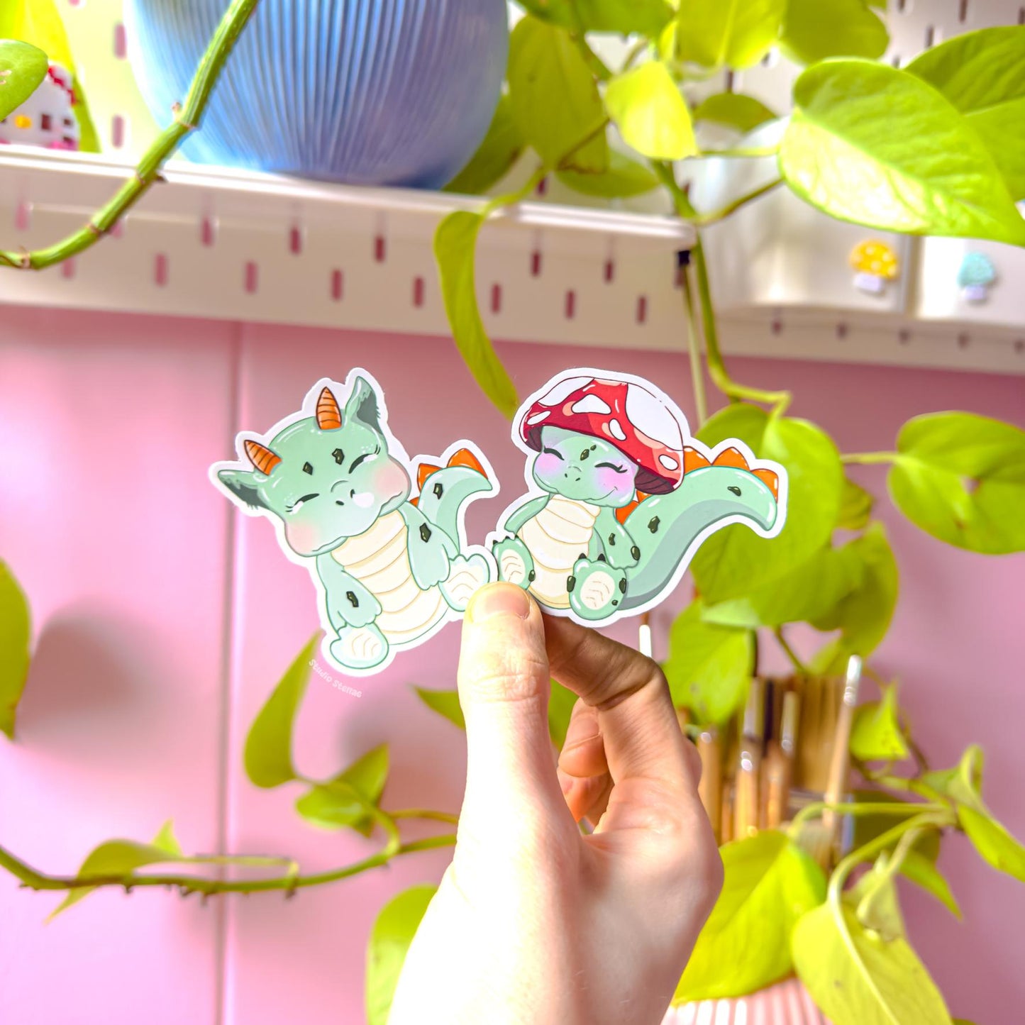 Lifestyle shot of a hand holding both dinosaur stickers near trailing green plants and soft daylight, demonstrating real-life scale and color accuracy. The cozy desk setting enhances the cottagecore aesthetic while showing how the decals look in everyday use. Perfect for personalizing planners, notebooks, laptops and water bottles with gentle pastel fantasy charm.