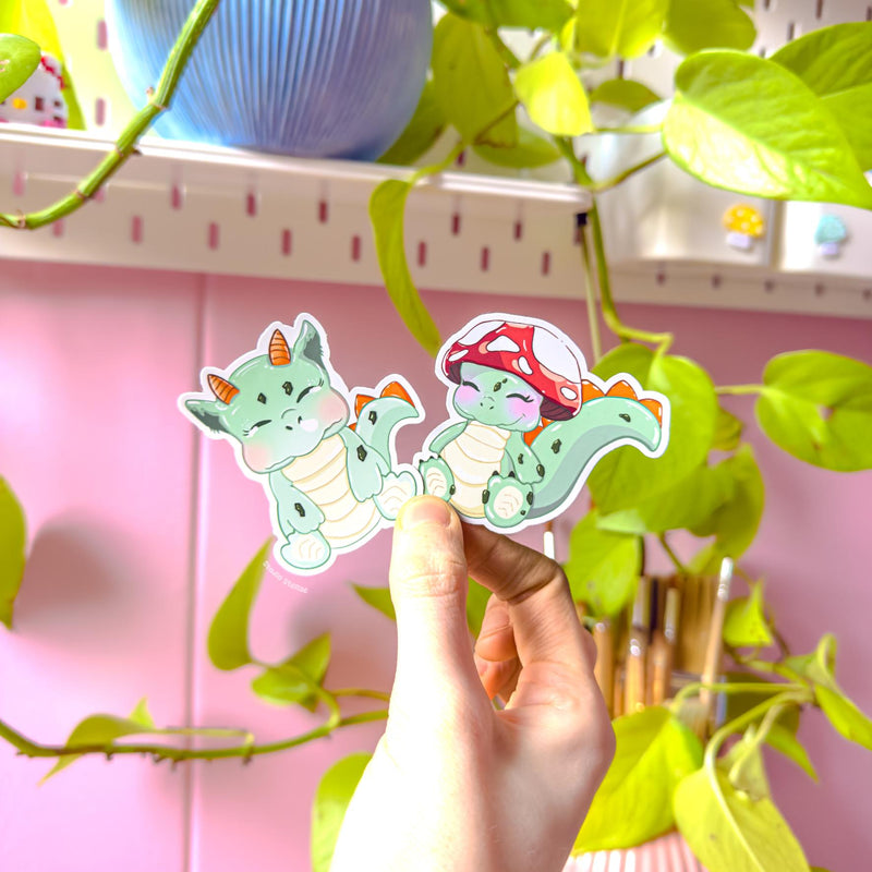 Lifestyle shot of a hand holding both dinosaur stickers near trailing green plants and soft daylight, demonstrating real-life scale and color accuracy. The cozy desk setting enhances the cottagecore aesthetic while showing how the decals look in everyday use. Perfect for personalizing planners, notebooks, laptops and water bottles with gentle pastel fantasy charm.