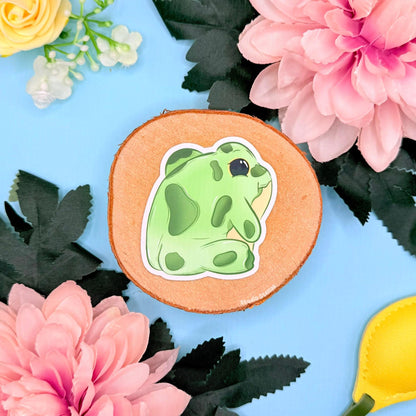 Flat lay of the frog bum sticker resting on cork with flowers and pastel props, showcasing its rounded shape and adorable little backside pose. The soft matte laminate and clean cut edges give it a premium handmade look. A charming green frog decal perfect for decorating planners, scrapbooks, laptops and bottles or gifting to frog fans and stationery lovers.