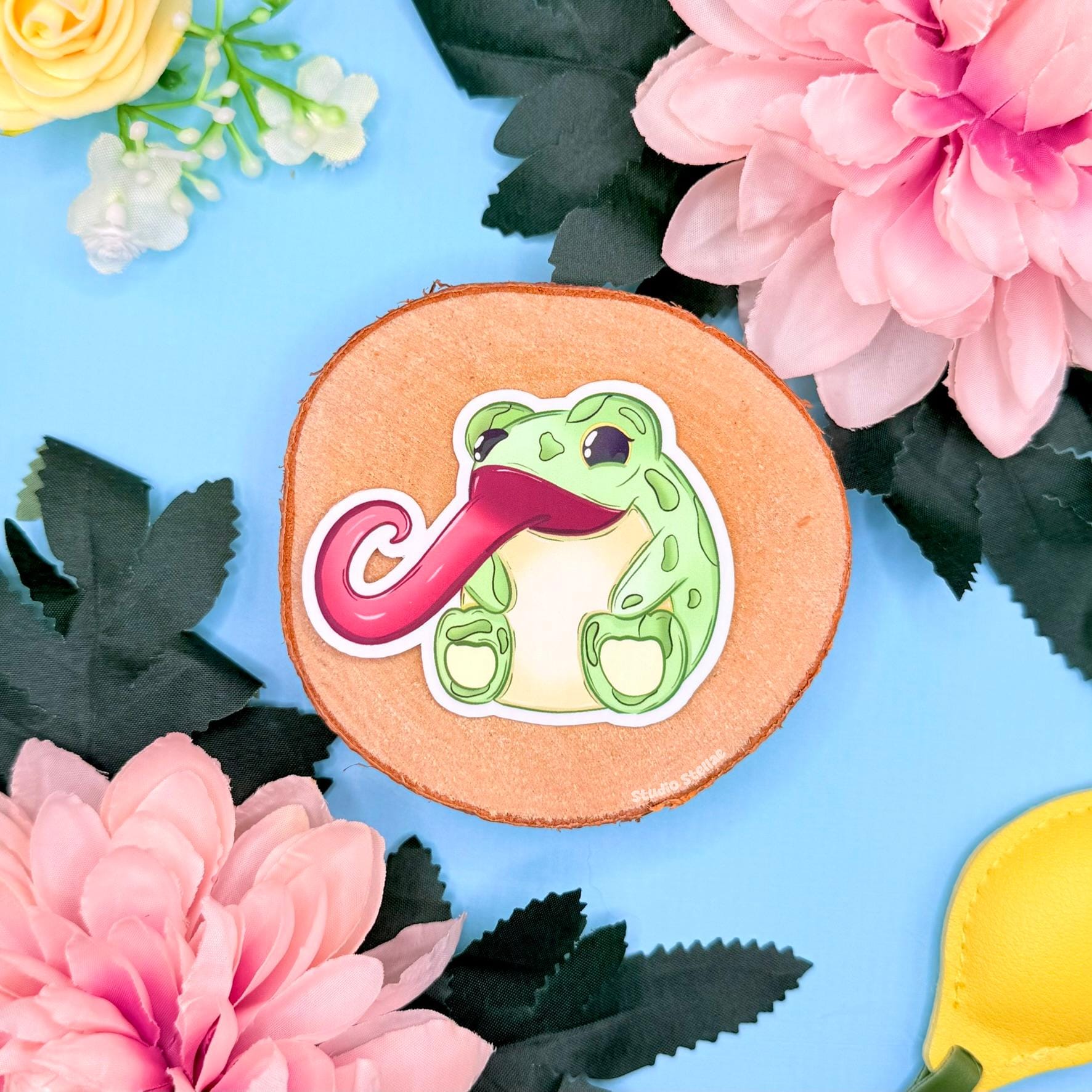 Flat lay of the long tongue frog sticker placed on cork with pink flowers and greenery, creating a bright playful scene that highlights the cheerful green color and smooth matte finish. This funny kawaii frog decal adds character to laptops, planners, journals and water bottles and makes a sweet gift for amphibian and cute animal lovers.