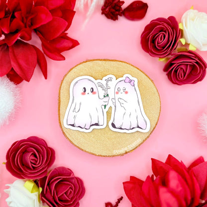 Two kawaii ghost couple stickers posed together on a cork board surrounded by red roses, with one ghost sweetly offering flowers to the other like a tiny romantic gift. The soft pastel pink background and floral props create a cozy Valentine and Halloween vibe, highlighting the cute spooky love story theme. Handmade, matte laminated, waterproof ghost decals designed for decorating planners, journals, laptops, water bottles or adding to a boo basket gift.