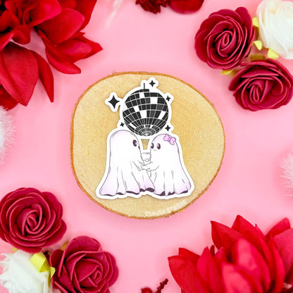 Close-up of two adorable ghost stickers dancing hand in hand on a cork surface framed by red roses, captured mid-twirl like a tiny spooky ballroom moment. Their soft white chibi shapes and gentle smiles give the scene a sweet romantic Halloween aesthetic with pastel tones. Handmade with a matte laminated, waterproof finish, these cute ghost couple decals are perfect for decorating planners, journals, laptops, water bottles or gifting to your favorite person.