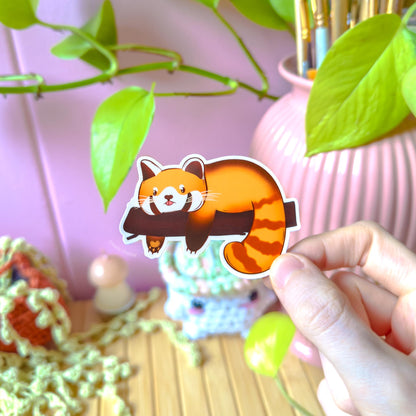 Hand holding the sleepy red panda sticker with a relaxed, curled-up pose that captures cozy, low-energy days. The soft colors and rounded chibi style give it a comforting feel, while the matte laminate reduces glare. This close-up emphasizes the “lazy mood” design, ideal for planners, notebooks or water bottles and relatable for anyone who loves cute animal humor and calm aesthetics.