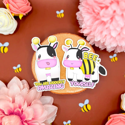 Cute kawaii cow dressed as a bumblebee sticker set featuring soft pastel colors, blushing cheeks, and a playful expression. One sticker reads “You are udderly amazing” and the other says “Bee yourself,” promoting self-acceptance, identity, and kindness. The stickers are styled on a warm-toned background with pink flowers and tiny bees around them. Perfect for decorating journals, laptops, planners, water bottles, or as an encouraging and inclusive gift that spreads positivity.