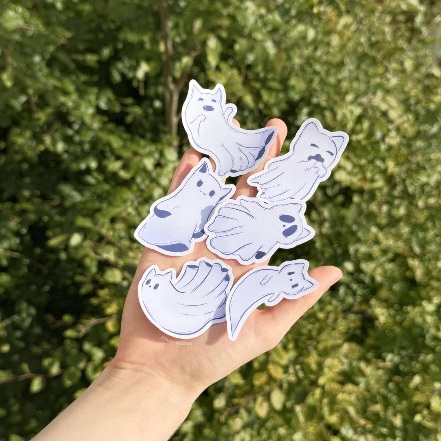 Hand holding the complete Ghost Kitty sticker set outdoors against fresh green grass and natural daylight, helping show true colors and real-life scale. The pastel ghost cats pop beautifully in the sunlight, making the soft purples, blues, and whites look vibrant and clean. This lifestyle photo demonstrates portability and everyday use while capturing the playful Halloween theme. Ideal for cat lovers decorating planners, bottles, laptops, or journals on the go.
