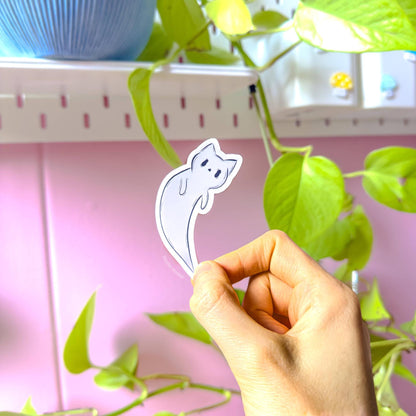 Hand holding an individual ghost kitty sticker up in front of leafy green houseplants and a soft pastel pink wall. The kawaii spooky cat floats like a little ghost, with a cute chibi expression and smooth white outline. This close-up shows the sticker’s real-life size, crisp die-cut edges, and vibrant pastel colors in natural light. Matte laminated, water-resistant, scratch-resistant handmade Halloween decal for planners, journals, laptops, and water bottles.