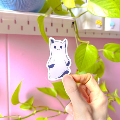 Hand-held close-up of an individual kawaii ghost kitty sticker presented against a bright, cozy studio corner with greenery and soft pink tones. This view clearly shows the full shape of the paranormal cat design, the neat white border, and the precise cut around the illustration. Natural light helps show true pastel colors and print clarity. Handmade matte laminated Halloween cat sticker, durable and water-resistant for planners, laptops, and bottle decor.