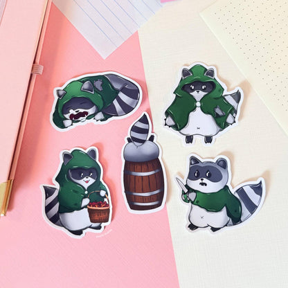 Rogue raccoon sticker set arranged on a soft pink and green background showing all characters clearly, including raccoon rogues with fantasy gear and potion bottle. This image highlights design variety and clean die cut edges, helping customers see details of the tabletop RPG sticker pack for decorating gaming accessories or stationery.