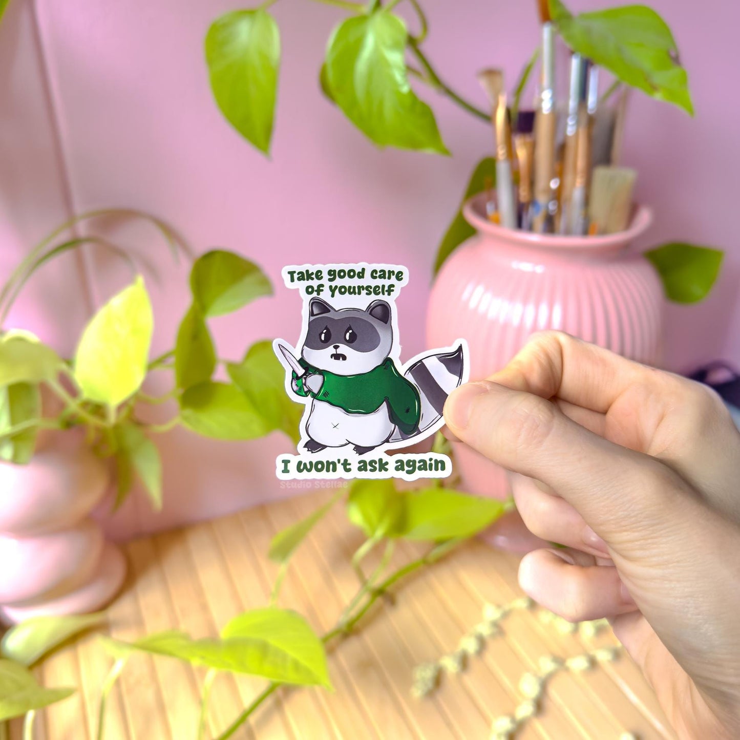 Hand holding the self care raccoon sticker near a pink desk and plants, showing real life scale, matte texture, and details of the cute trash panda design with knife and quote. This lifestyle photo demonstrates how the funny mental health sticker looks when applied to everyday surfaces like planners, laptops, or water bottles, highlighting its playful sarcastic humor and handmade quality.