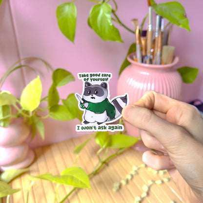 Hand holding the self care raccoon sticker near a pink desk and plants, showing real life scale, matte texture, and details of the cute trash panda design with knife and quote. This lifestyle photo demonstrates how the funny mental health sticker looks when applied to everyday surfaces like planners, laptops, or water bottles, highlighting its playful sarcastic humor and handmade quality.