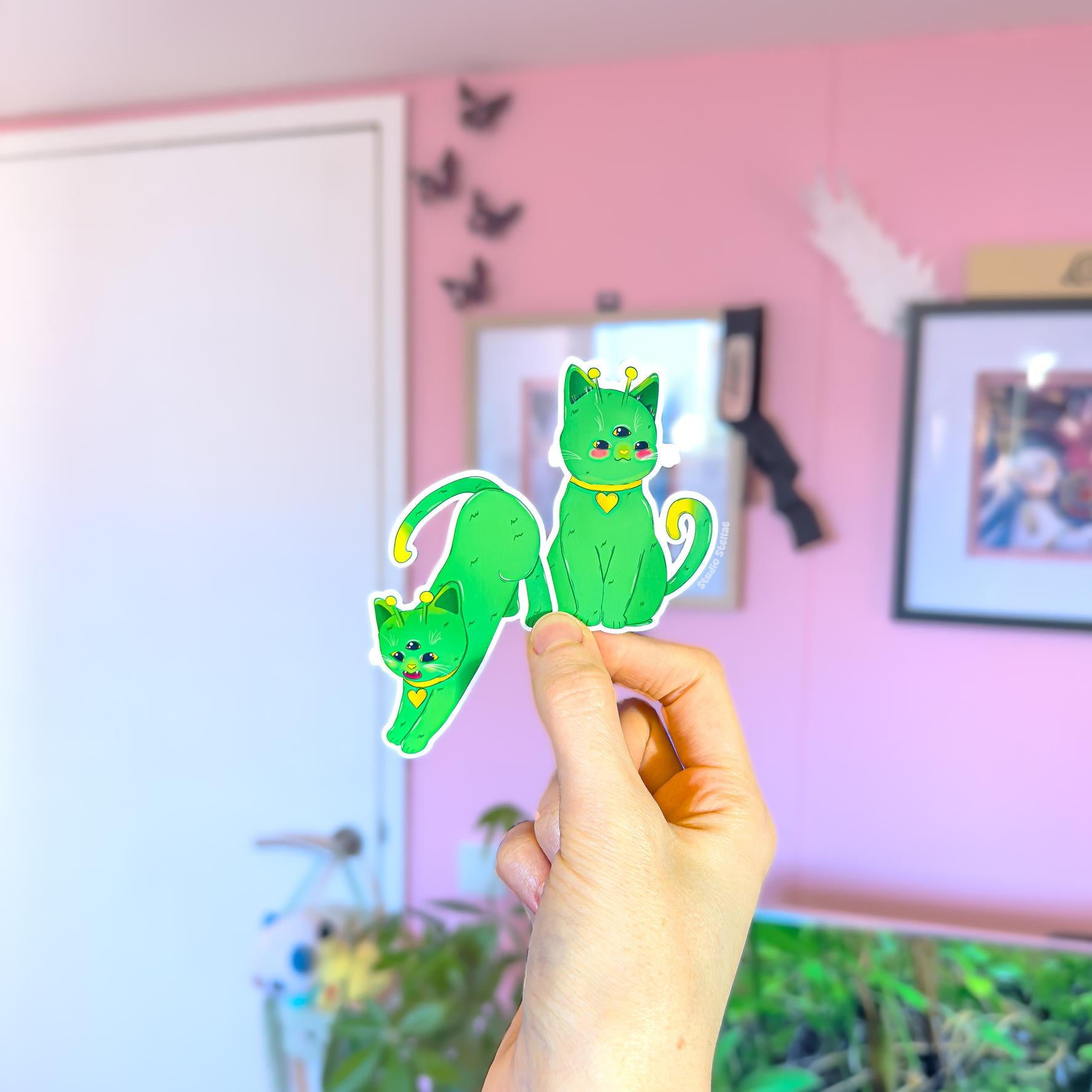 Hand holding an alien cat sticker against a pink room background, showing real life scale, bright colors, and matte finish of the green space kitty design. This lifestyle image demonstrates how the cosmic cat decal looks when used on planners, laptops, or water bottles, highlighting the playful sci fi theme and cute chibi style that makes this sticker set perfect for cat lovers and fans of quirky outer space aesthetics.