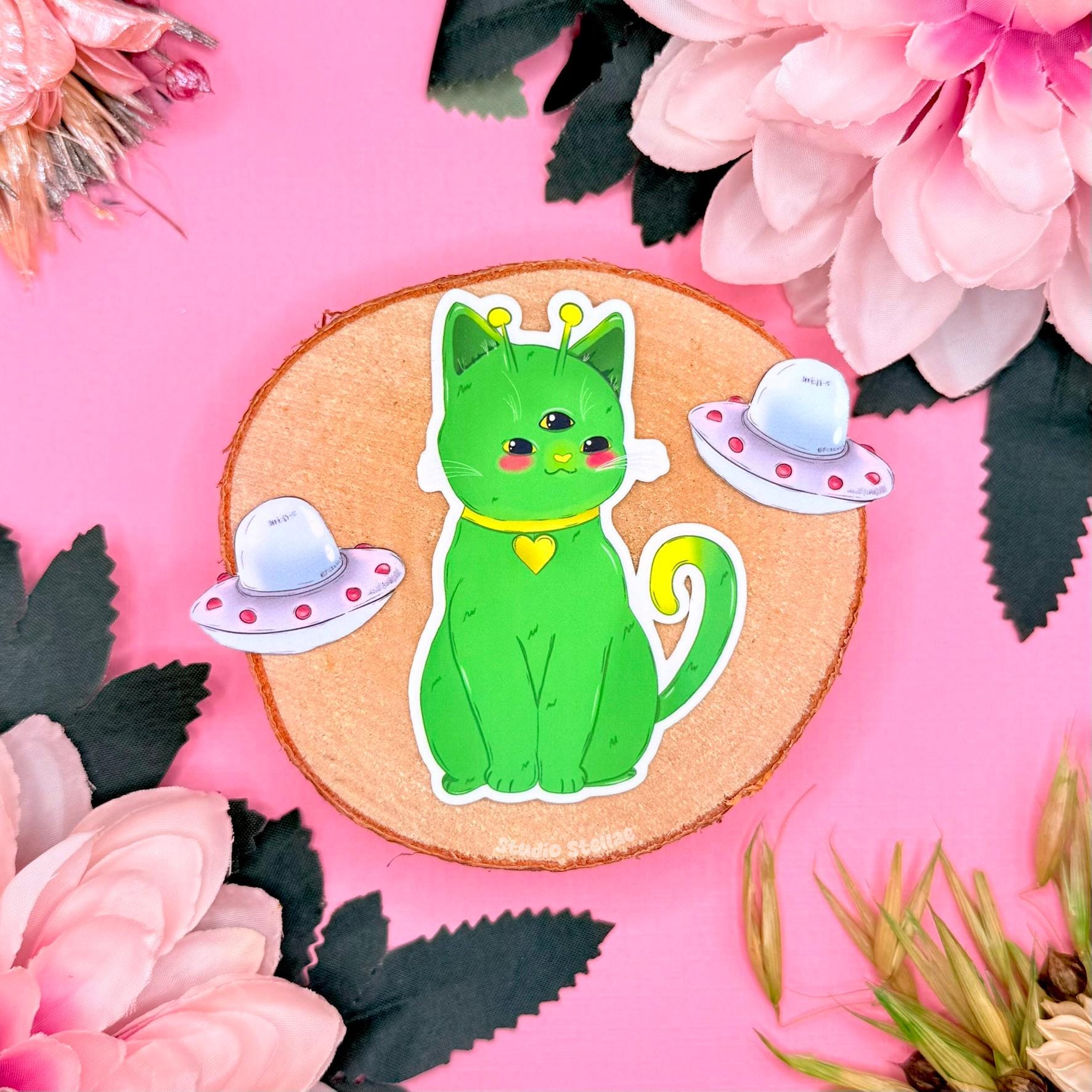Alien cat sticker set close-up on a pink floral background, featuring the green space kitty sitting upright with tiny pastel UFO props around it. This photo highlights the cute chibi alien cat face, smooth matte laminated finish, and clean die cut edge. A whimsical sci fi cat sticker perfect for decorating planners, journals, laptops, and water bottles, ideal for cat lovers who collect kawaii cosmic trinkets and outer space themed stationery.