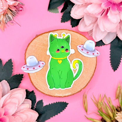 Alien cat sticker set close-up on a pink floral background, featuring the green space kitty sitting upright with tiny pastel UFO props around it. This photo highlights the cute chibi alien cat face, smooth matte laminated finish, and clean die cut edge. A whimsical sci fi cat sticker perfect for decorating planners, journals, laptops, and water bottles, ideal for cat lovers who collect kawaii cosmic trinkets and outer space themed stationery.