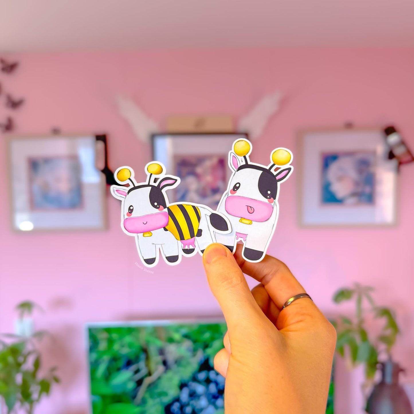 Hand holding a bee cow sticker in a cozy pink room, showing real life scale and the cute chibi details—pink nose, little wings, and striped bee outfit. The matte laminated finish and clean outline are visible, helping customers imagine the sticker on a laptop, planner, water bottle, or phone case. A sweet nostalgic gamer stationery gift for MySims Kingdom fans and anyone who loves soft farm game vibes.