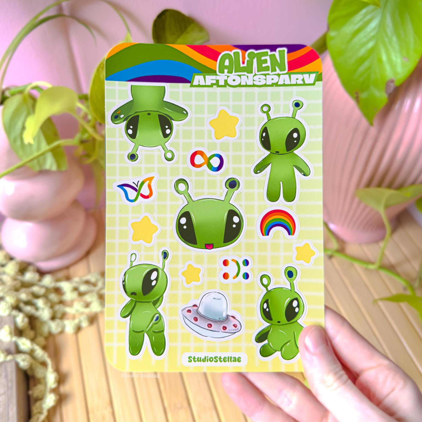 Hand holding the Aftonsparv alien sticker sheet near a cozy plant setup, showing real life scale and how the colorful alien designs look in natural light. The soft green palette and cute neurodivergent icons stand out clearly, helping customers imagine using these IKEA alien stickers for decorating planners, laptops, iPads, or journals while expressing ADHD pride, autism acceptance, and love for kawaii stationery.