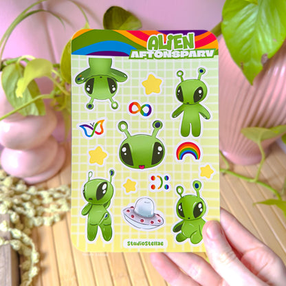 Hand holding the Aftonsparv alien sticker sheet near a cozy plant setup, showing real life scale and how the colorful alien designs look in natural light. The soft green palette and cute neurodivergent icons stand out clearly, helping customers imagine using these IKEA alien stickers for decorating planners, laptops, iPads, or journals while expressing ADHD pride, autism acceptance, and love for kawaii stationery.