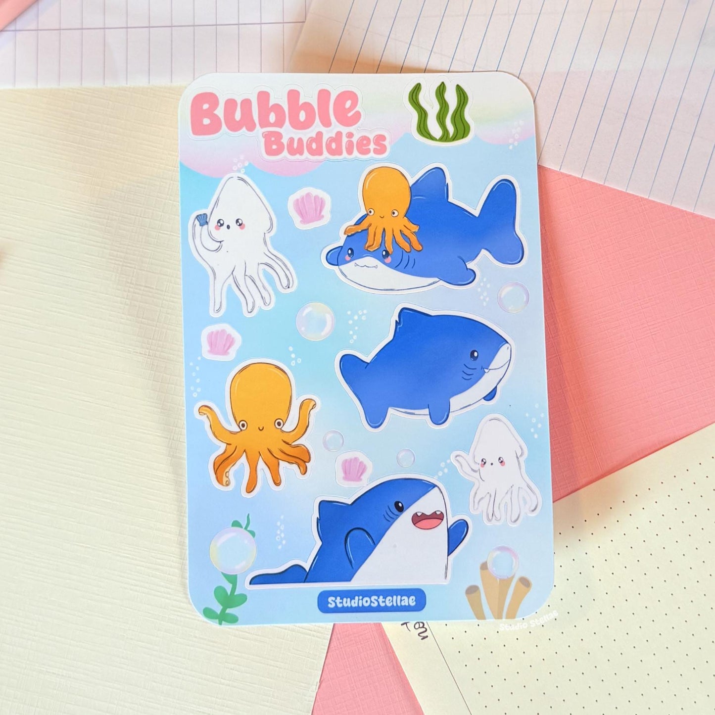 Sticker sheet photographed against a simple background indoors, focusing on the artwork without distractions. This clean view allows shoppers to easily see the individual shark, squid, octopus, bubble, and shell stickers and understand the overall composition of the sheet.