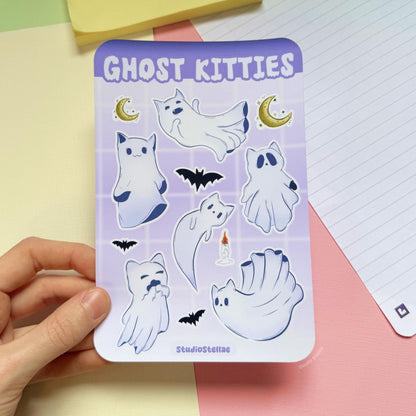 Hand holding the Ghost Kitties sticker sheet above a pastel desk with a notebook in the background. The full sheet shows white ghost cats with soft purple outlines, crescent moons, and small bats in a pastel goth Halloween style. This lifestyle image helps show real-life scale and size in hand, perfect for journaling, planner decorating, scrapbooking, laptops, and adding spooky witchy vibes to everyday surfaces.