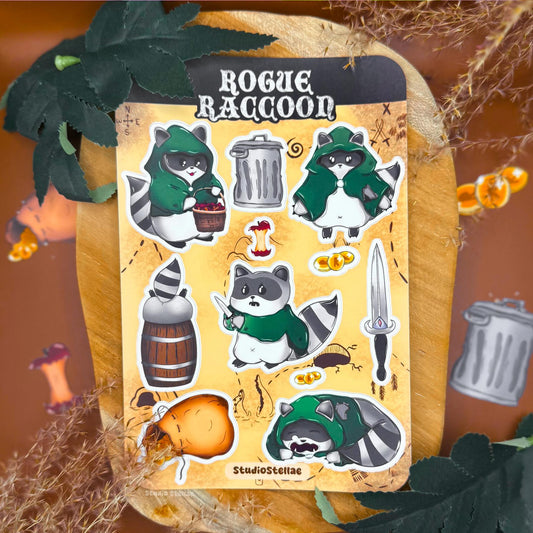 Rogue Raccoon sticker sheet styled in a cozy fantasy scene with autumn leaves, warm lighting, and soft textures. The sheet shows chibi raccoon rogue characters wearing green cloaks, holding loot and daggers, alongside accessories like gold coins, a trash can, barrel, treasure sack, and fantasy props. The matte laminated surface and rounded corners are visible, creating a cute DnD and tabletop RPG inspired aesthetic perfect for raccoon lovers and fantasy sticker collectors.