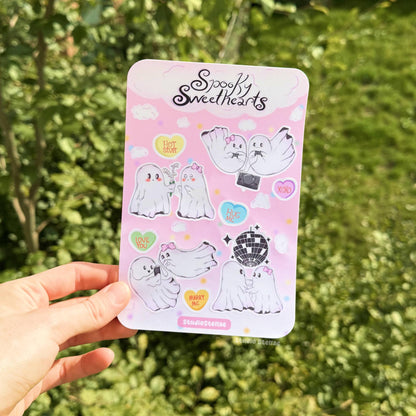 Hand holding the Spooky Sweethearts sticker sheet outdoors against a lush green grass background in natural daylight. The full sheet shows romantic ghost couples in sweet scenes like dancing, gaming, and sharing moments together, along with candy hearts and soft cloud stickers. This lifestyle photo demonstrates real-life scale, color clarity, and how the stickers look in natural light, perfect for Valentine decorating, journaling, planners, or adding cute spooky romance to everyday items.