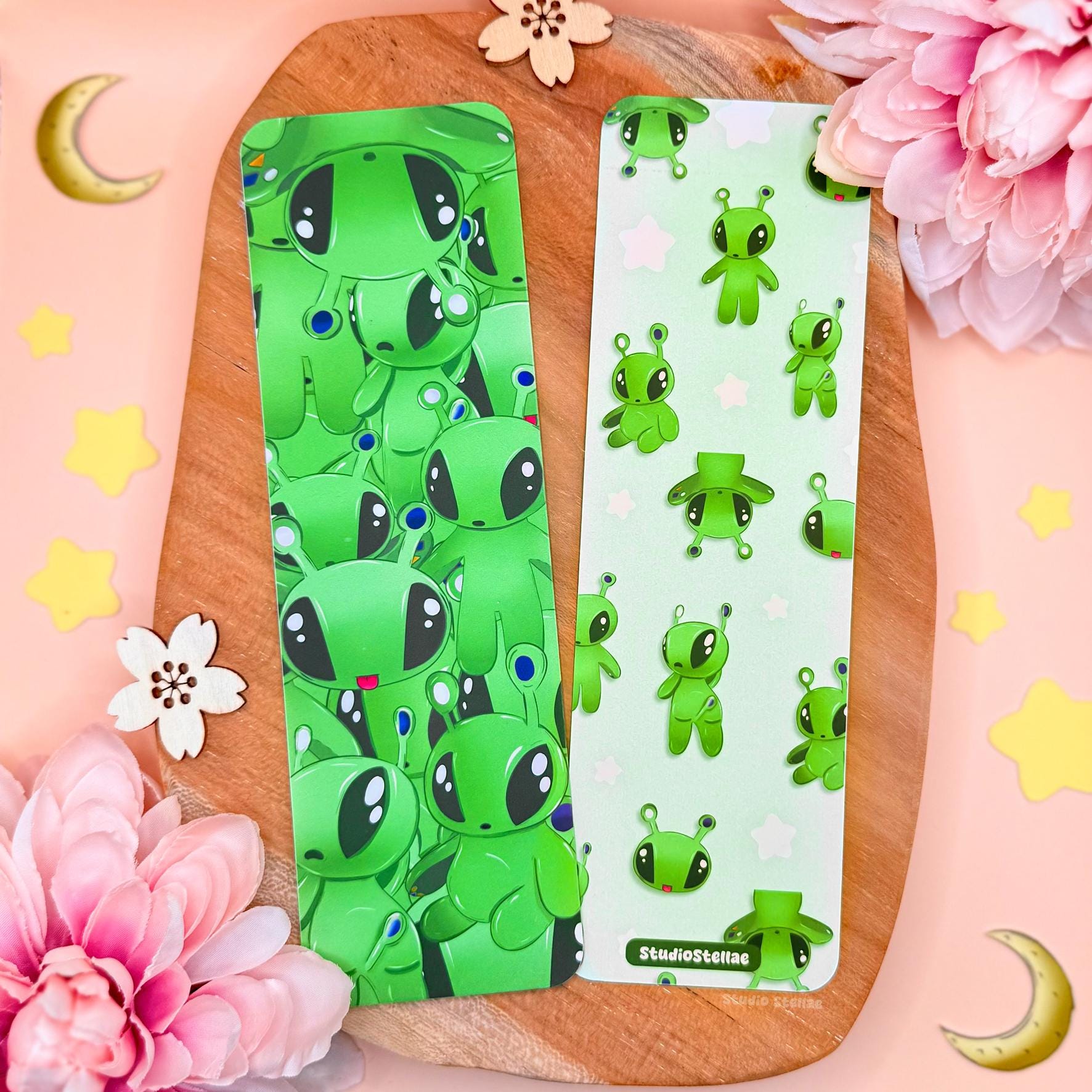 Two Aftonsparv-inspired green alien bookmarks displayed on a wooden tray with soft pink flowers and pastel props. The bookmarks feature cute plushie-style alien faces in bright green with big black eyes and tiny antenna details. One bookmark has an all-over alien pattern, the other shows a clustered alien print. The matte laminated finish and rounded corners are visible, creating a playful space-themed book accessory and reader gift aesthetic.
