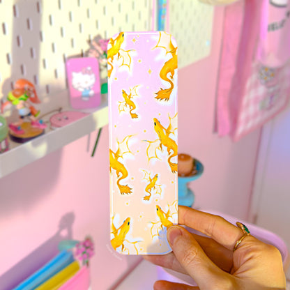 Hand holding the pastel gold dragon bookmark in front of a cozy decorated reading nook with soft pink and pastel accents in the background. The golden dragon design, clouds, and dreamy star details are clearly visible, showing how the bookmark looks in real-life use. This lifestyle photo emphasizes the romantasy vibe and makes it easy to imagine the bookmark tucked into fantasy novels, BookTok reads, journals, or planners as a cute laminated page marker.