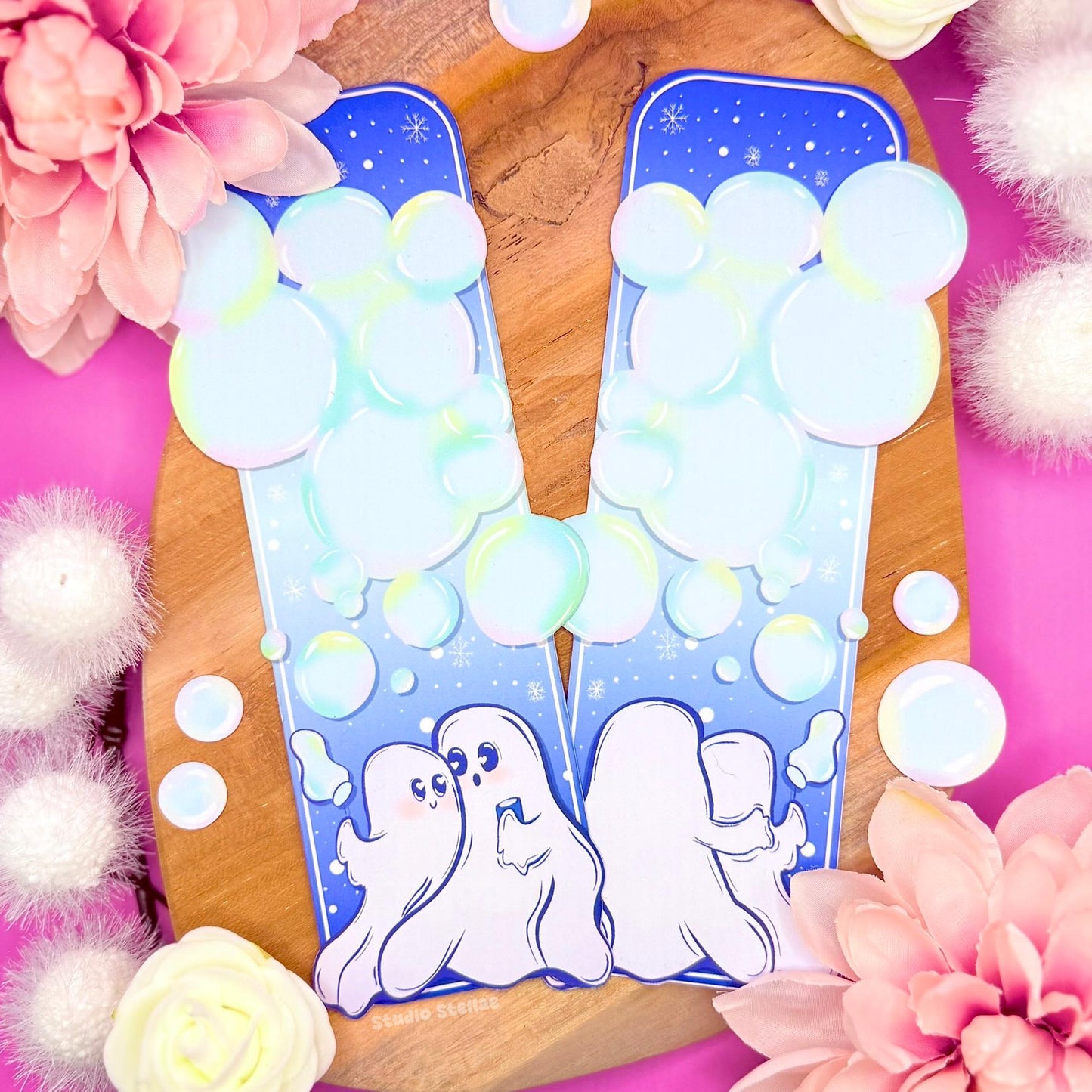 Ghost Couple Blowing Bubbles Bookmark displayed upright against a soft pink floral background. The uniquely shaped pastel bookmark features two romantic white ghosts at the bottom, blowing translucent blue bubbles beneath a starry night sky. The dreamy winter color palette blends soft blues and mint tones with a matte laminated finish. This romantic spooky page marker is perfect for Halloween gifts, Valentine gifts, and cozy reading aesthetics.