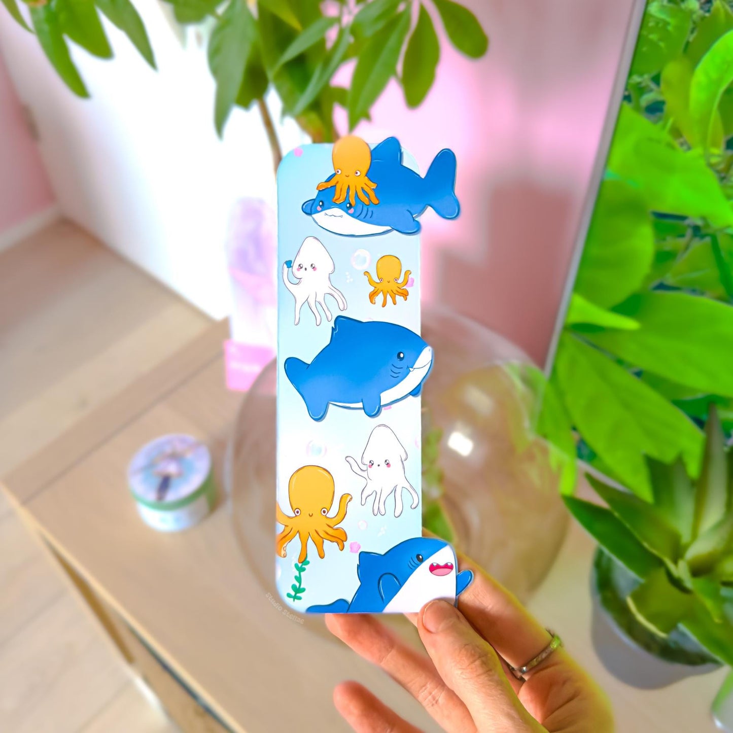 Hand holding the Pastel Shark Bookmark in a cozy workspace with a pink wall and leafy green plant in the background. The ocean animal page marker shows chibi sharks, octopus, and squid swimming through soft blue water. This lifestyle photo demonstrates real-life scale, thickness, and how the double-sided matte laminated bookmark looks in hand for readers and stationery lovers.